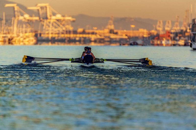 A single sculler on calm water at first light, with port cranes behind